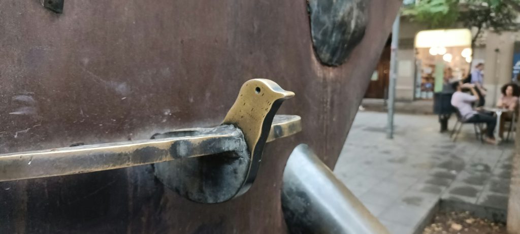Petite photo du quartier de Gracia, on y voit un petit oiseau métallique sur une aire de jeu avec en fond des personnes assises au bar