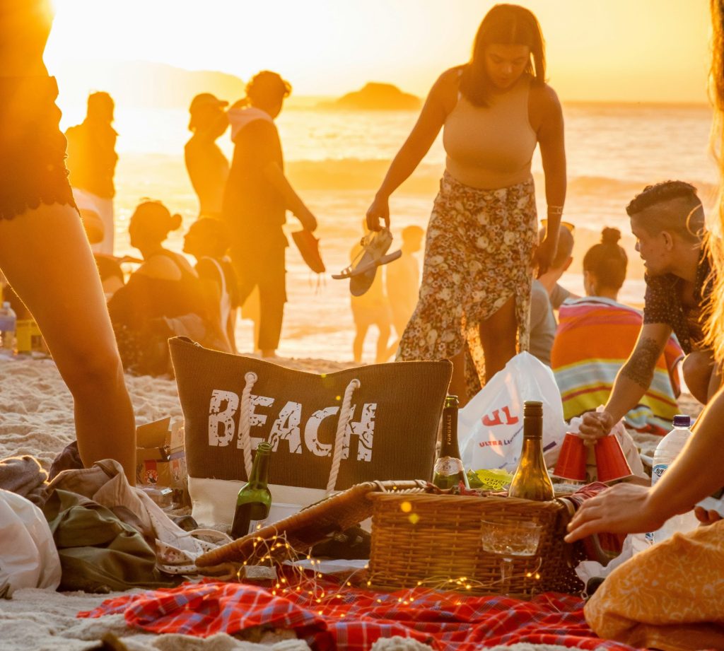 Beaucoup de personnes sur une plage au coucher de soleil. Les sacs et paniers repas sont de sortis avec bières, vins et apéritifs pour faire la fête dans la soirée