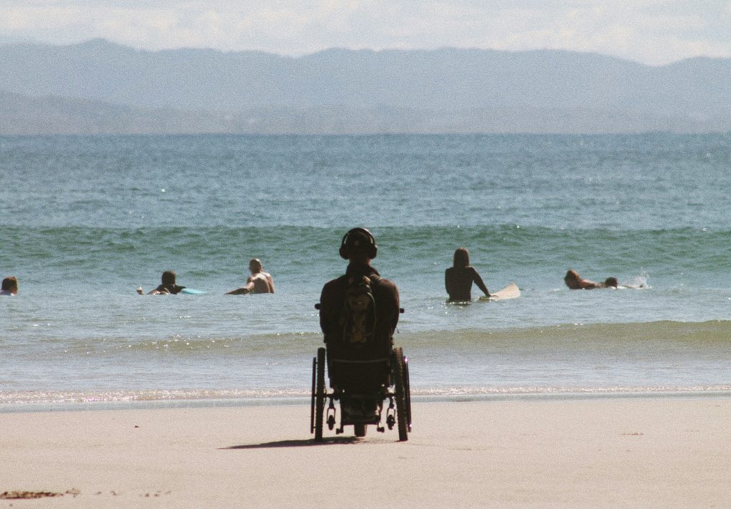 Une personne en fauteuil roulant à la plage qui observe, dos à la caméra, les nageurs et surfeurs dans l'eau